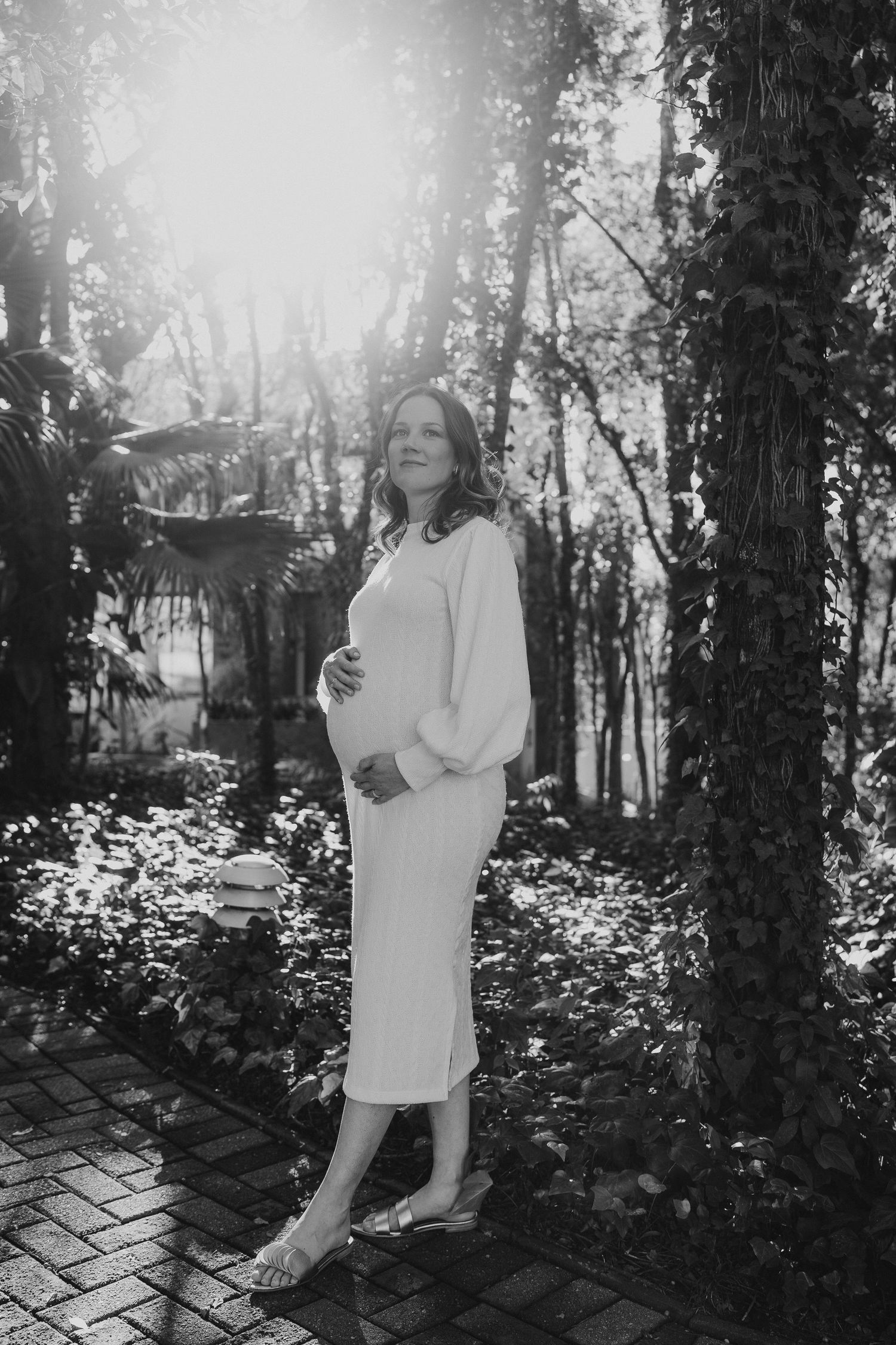 Serene maternity portrait in white dress amid sunlit forest setting.