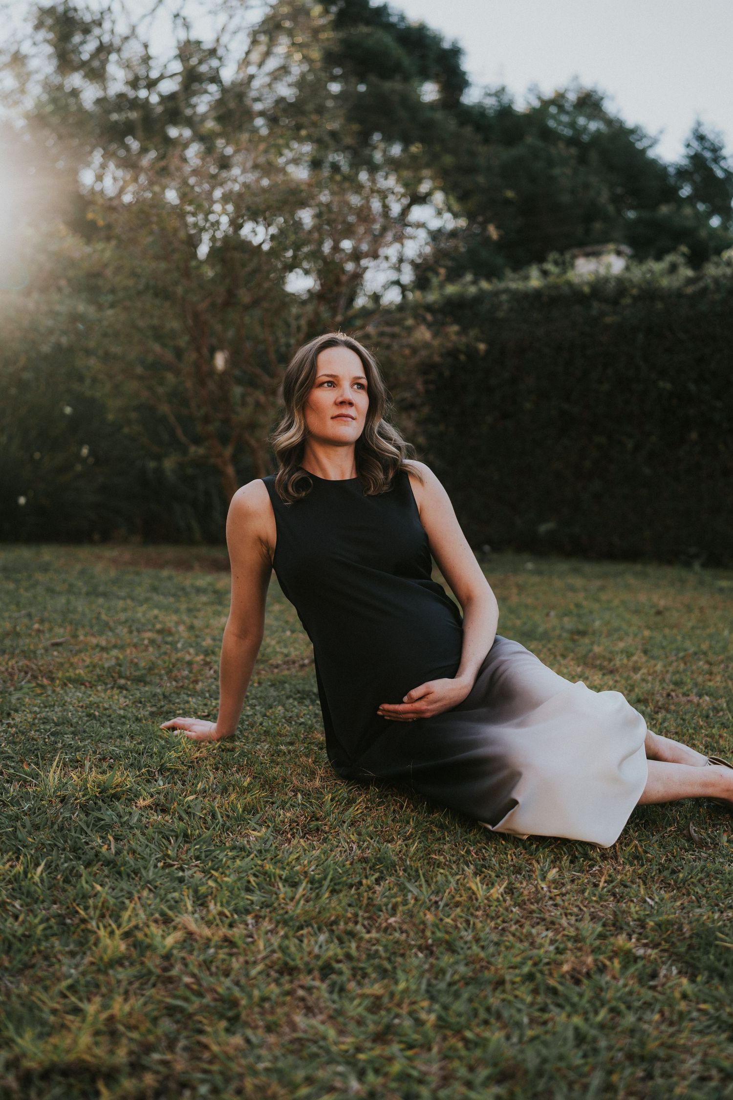 Maternity portrait of someone in a black dress reclining on grass at sunset.