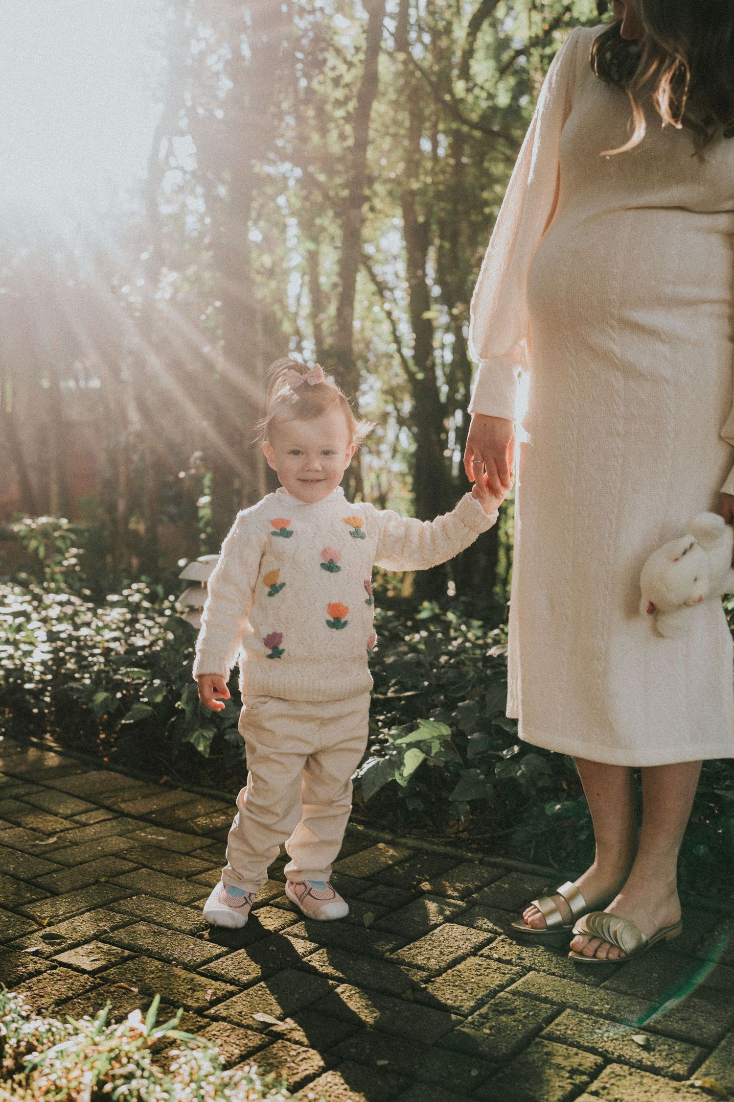 A small child in a cream outfit holds hands with an adult on a brick path surrounded by sunlit trees.