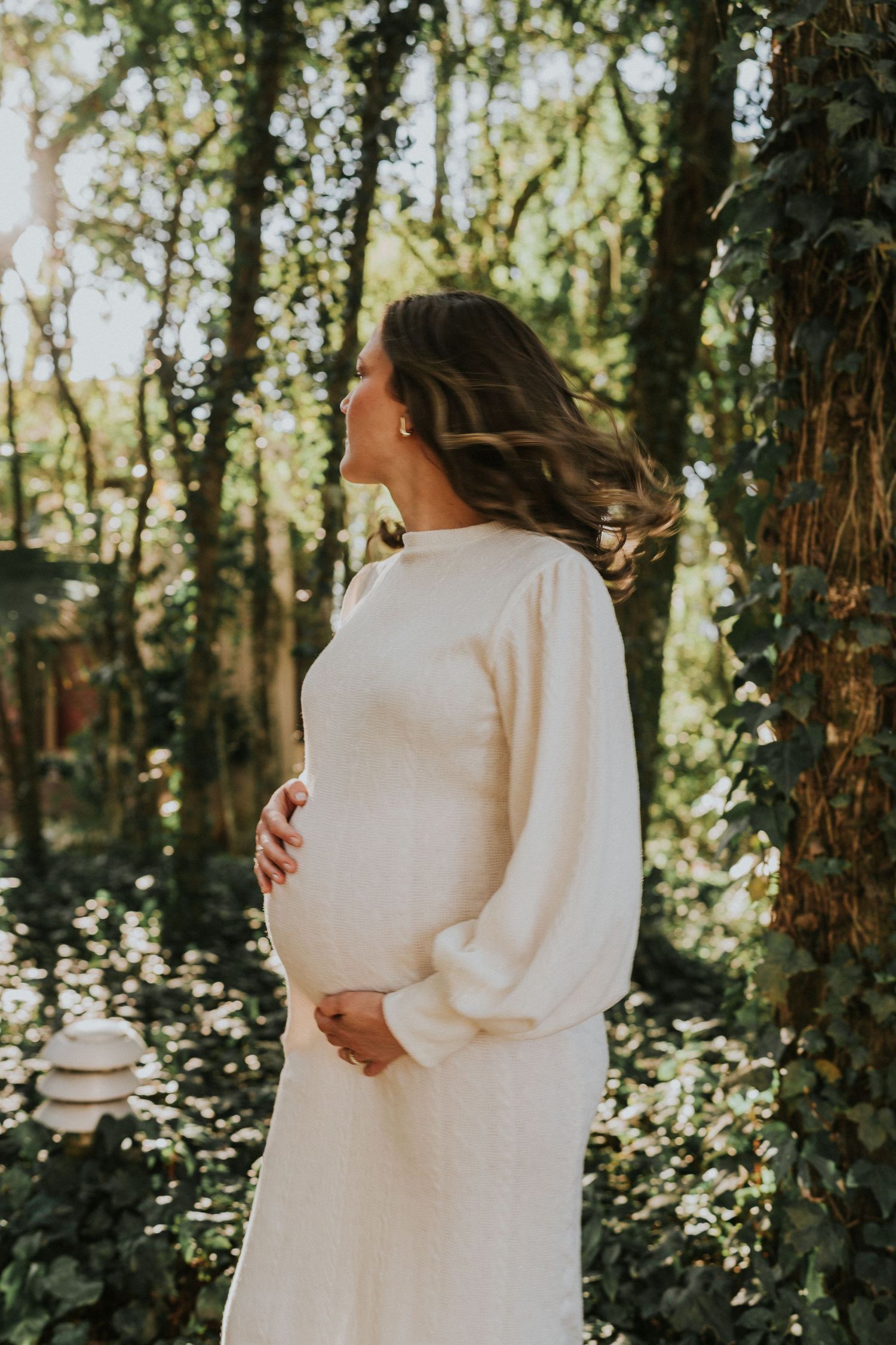 White-dressed figure with flowing hair poses in a sunlit forest setting during a maternity photoshoot.
