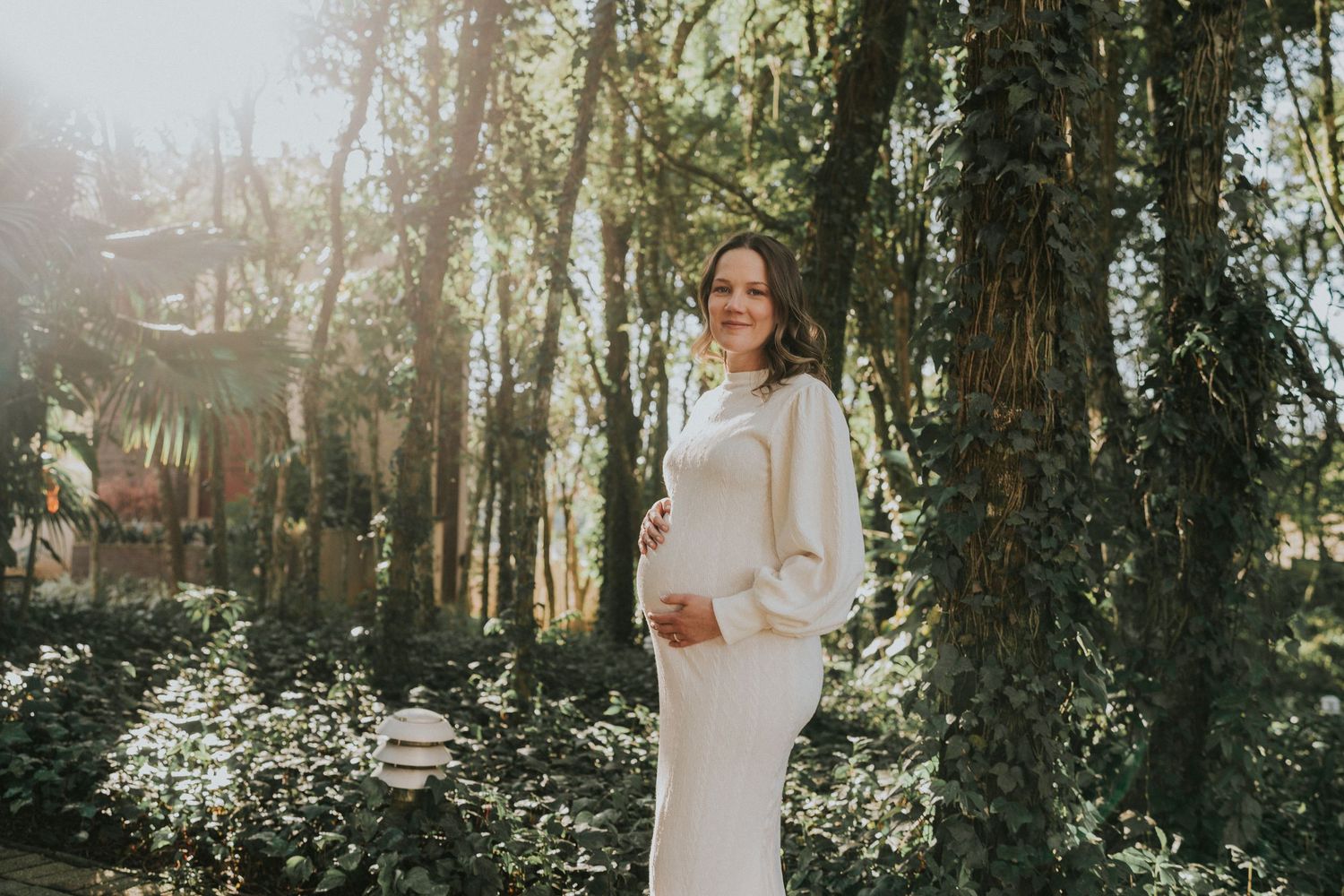 Elegant maternity photo in a white dress among sunlit trees and foliage.