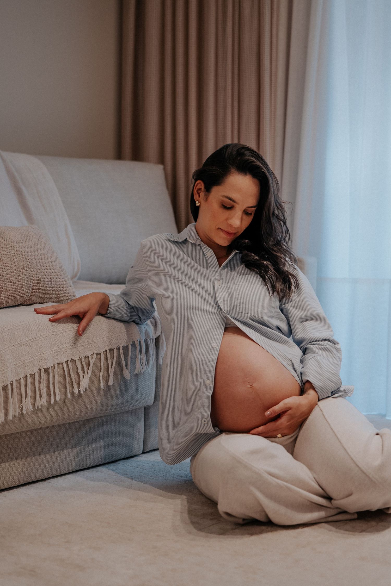 Maternity photos in soft lighting showing gentle moments on a couch with grey clothing.