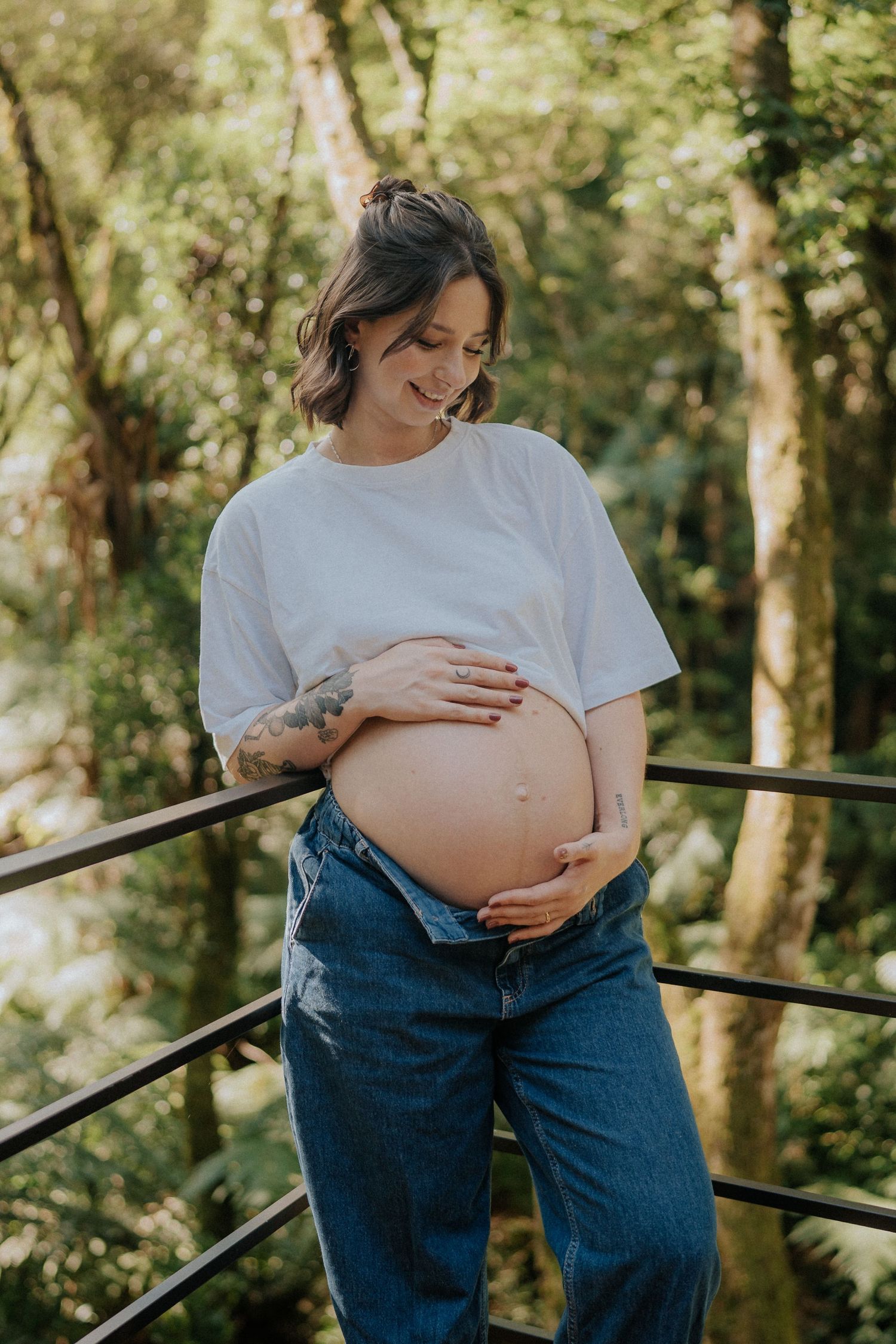 Expecting mother in white tee and jeans poses in sunlit forest setting.