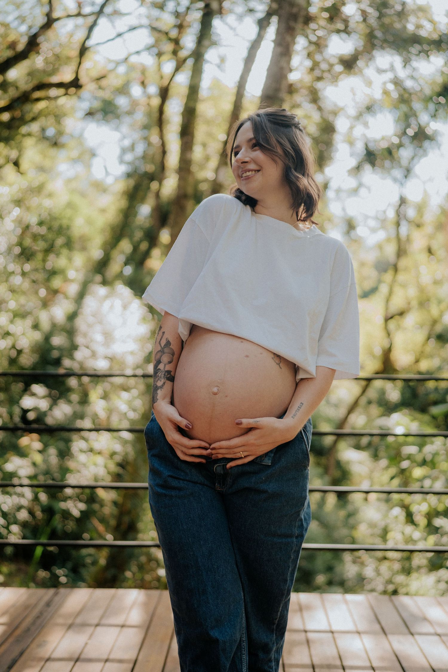 Expectant mother in white top poses against forest backdrop.