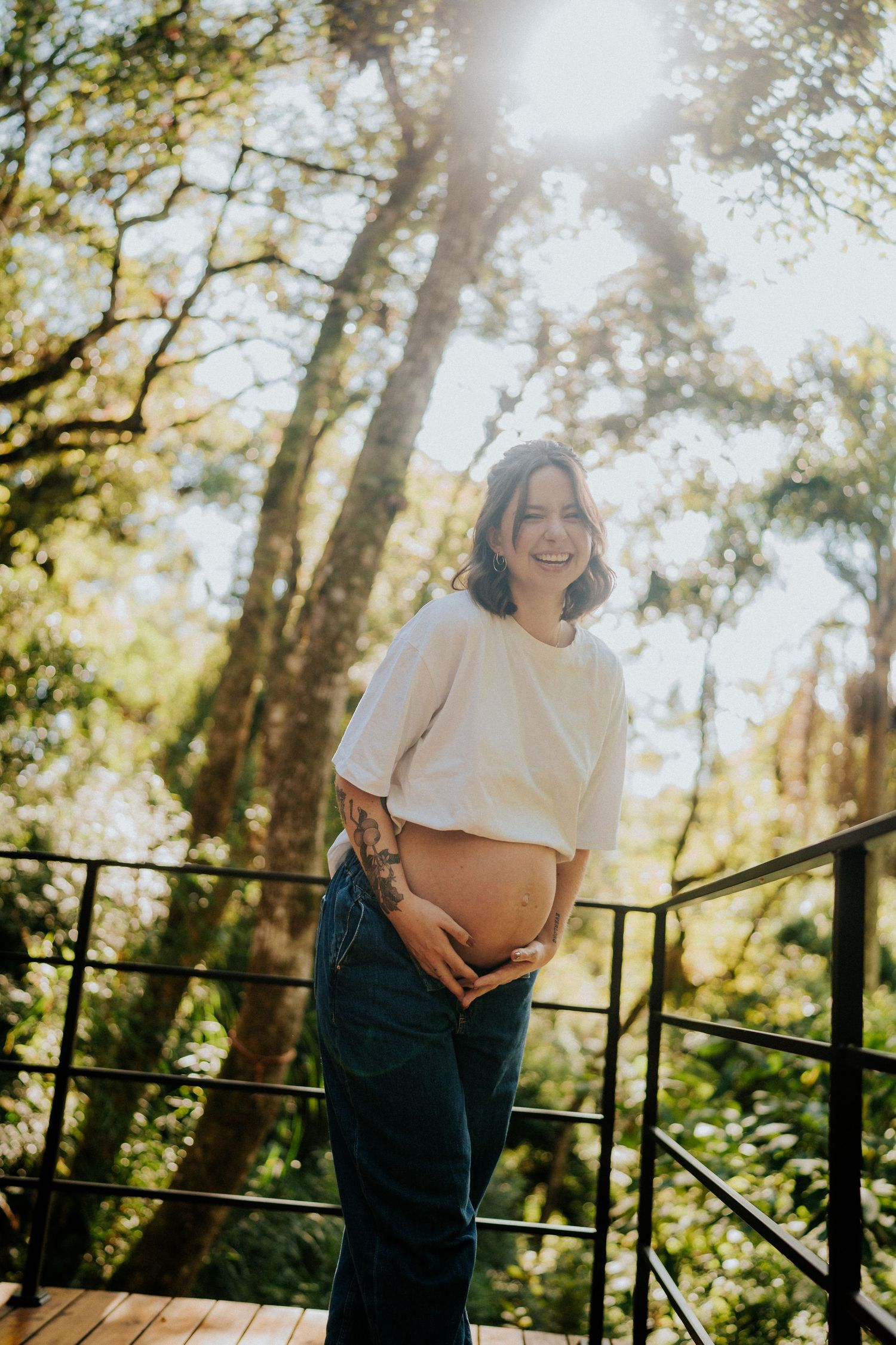Sun-drenched maternity portrait on wooden deck with forest scenery.