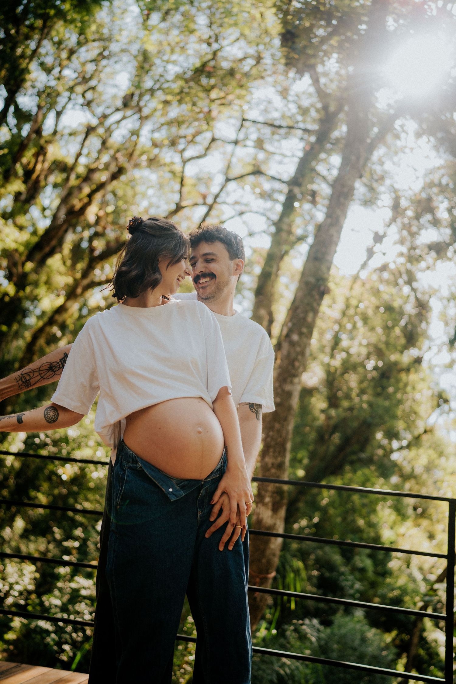 Intimate maternity photoshoot outdoors showing expectant couple embracing in dappled sunlight on wooden deck.