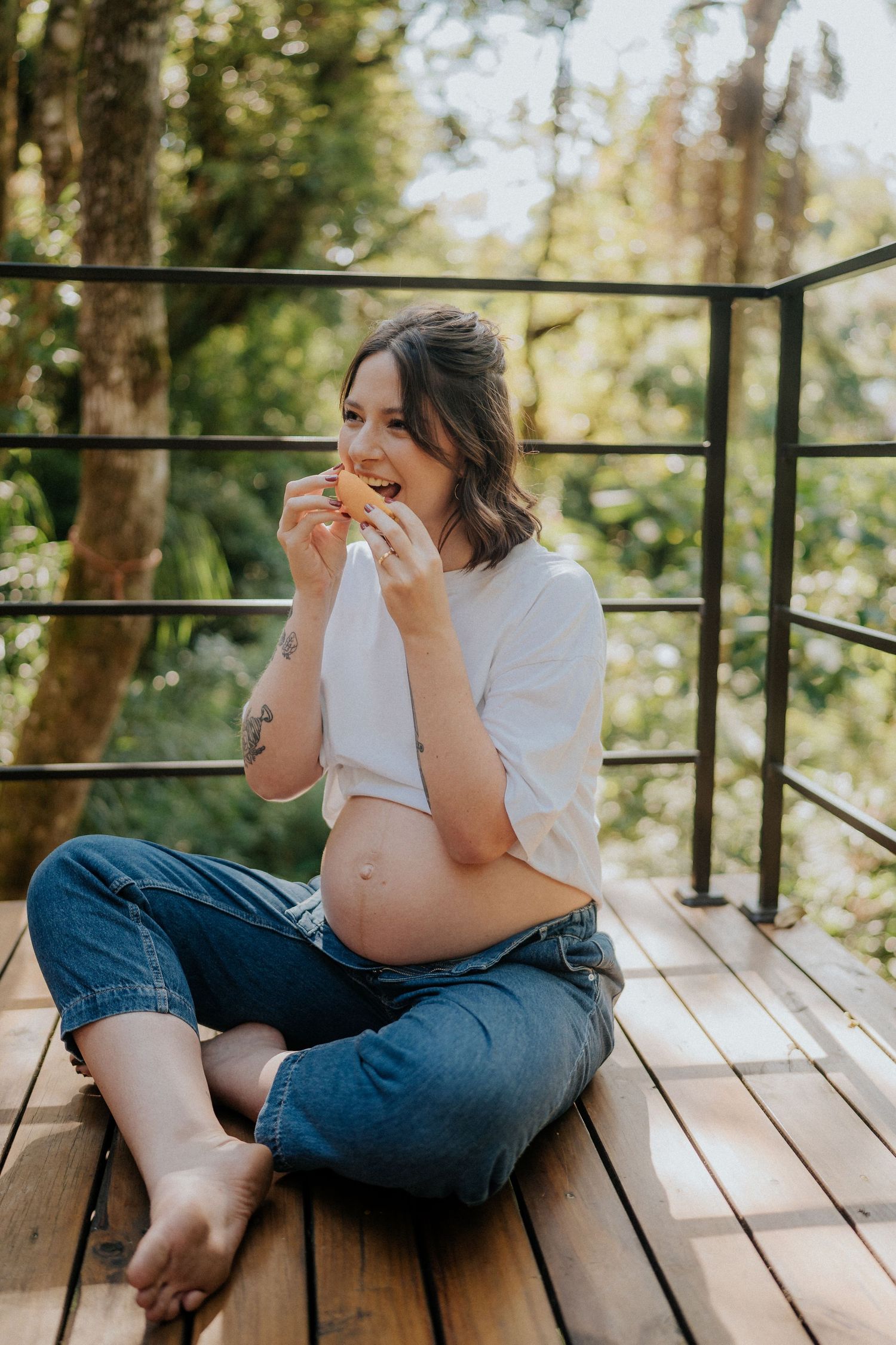 Expecting mother in white shirt and jeans seated on wooden deck enjoying a snack outdoors.