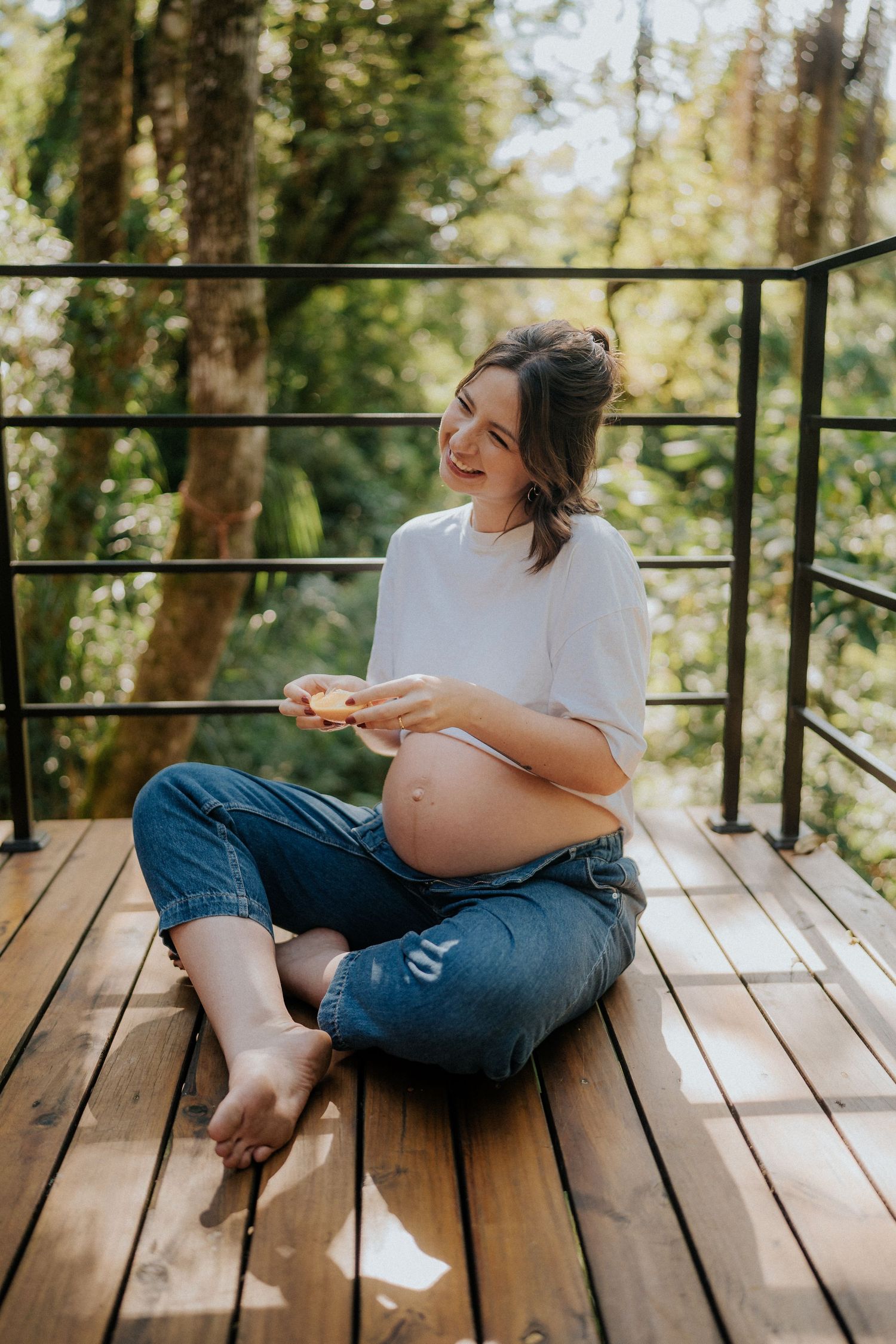 Pregnancy photography on wooden deck showing expecting mother in casual attire among trees.