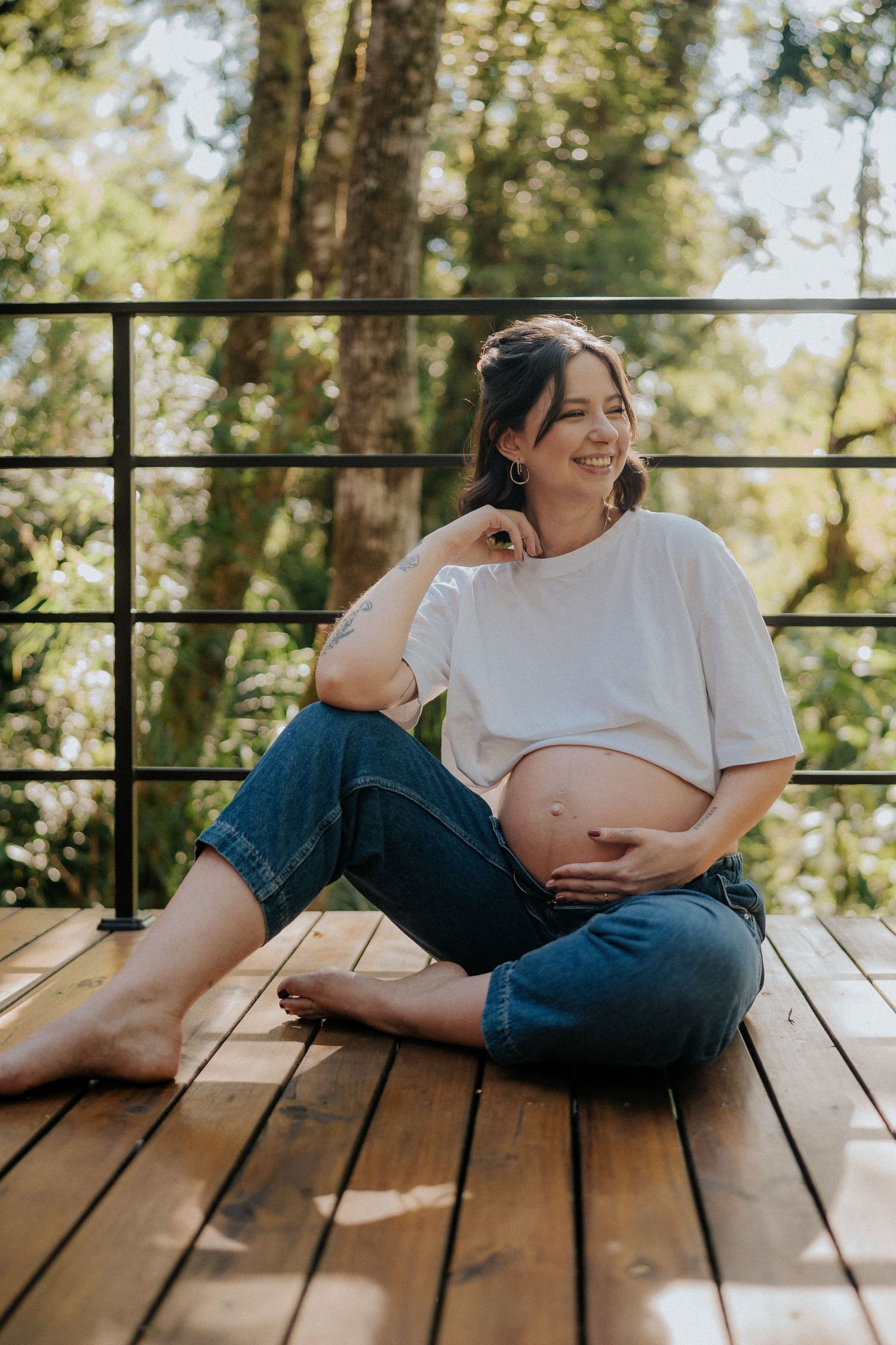 Candid maternity photo of expectant mother sitting cross-legged on wooden deck in natural setting.