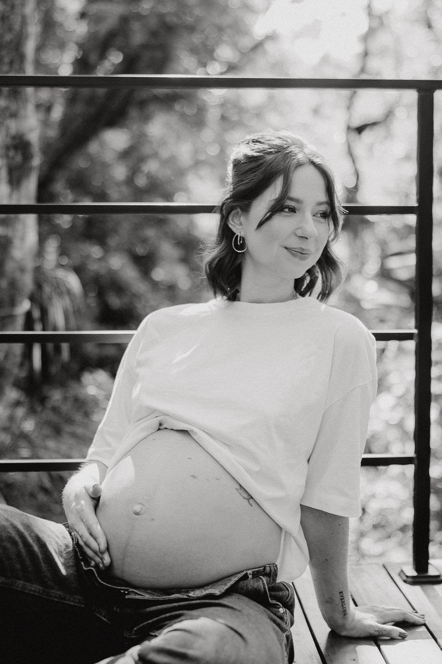 Black and white maternity portrait showing pregnant woman in white shirt sitting on outdoor deck.