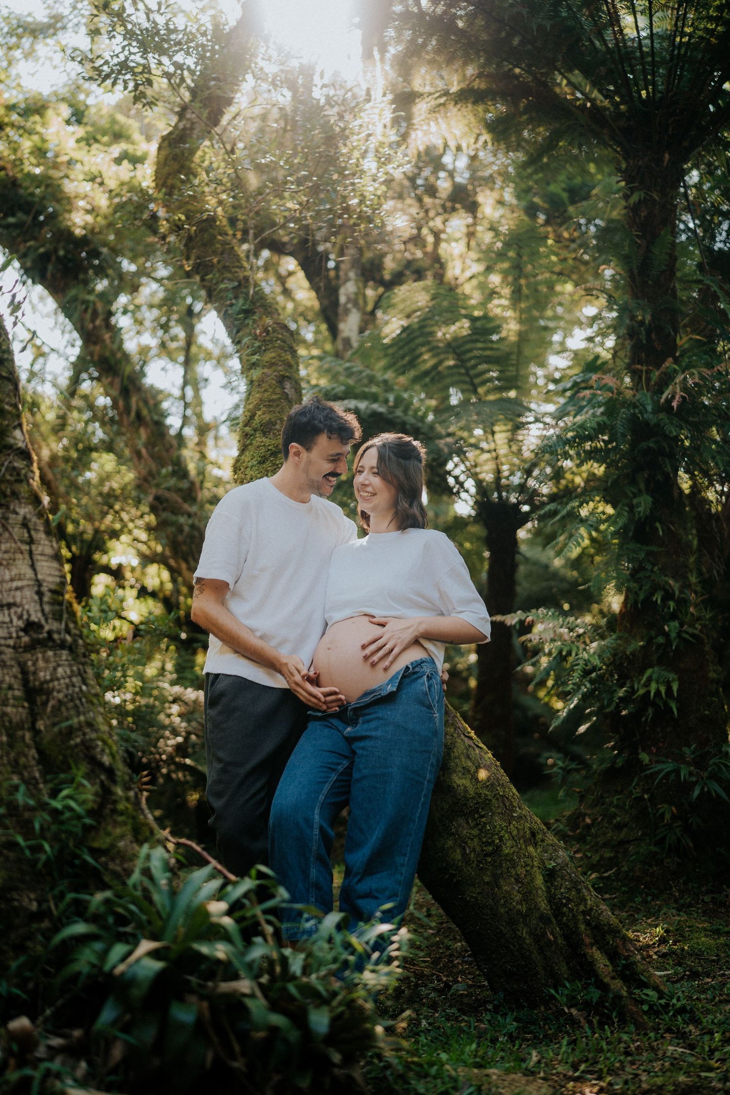 Sun-dappled forest maternity photo showing couple sharing intimate moment among tropical foliage.