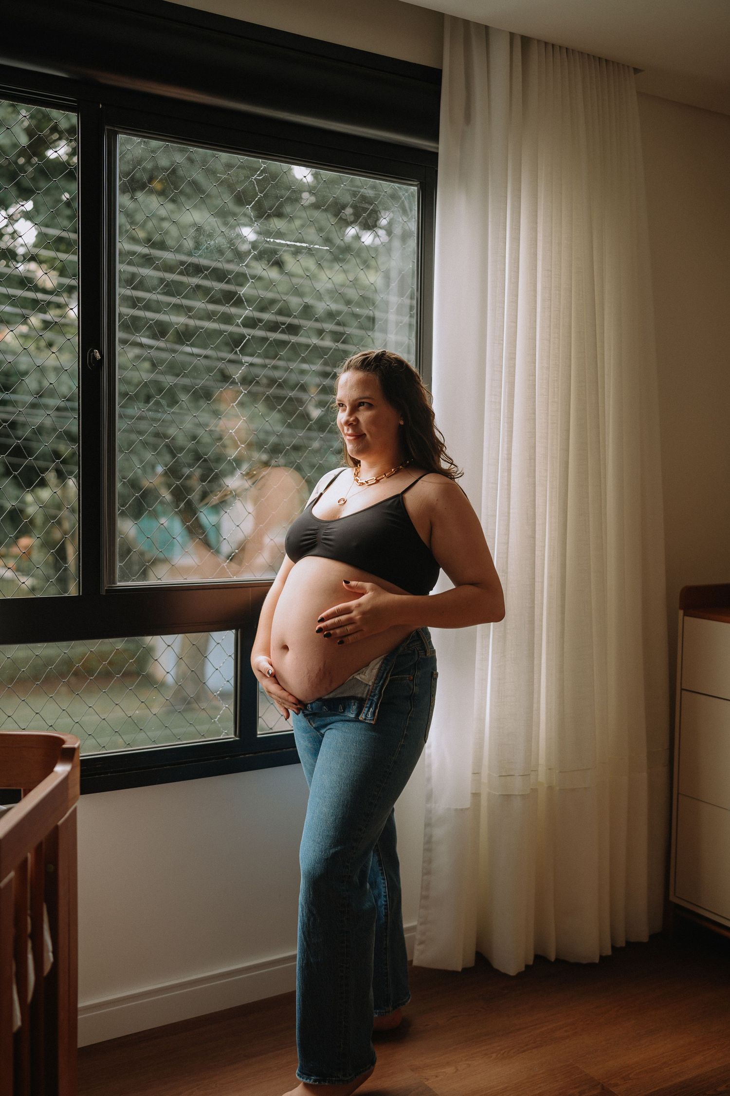 Casual maternity portrait of expecting mother in jeans by window showing bare belly.