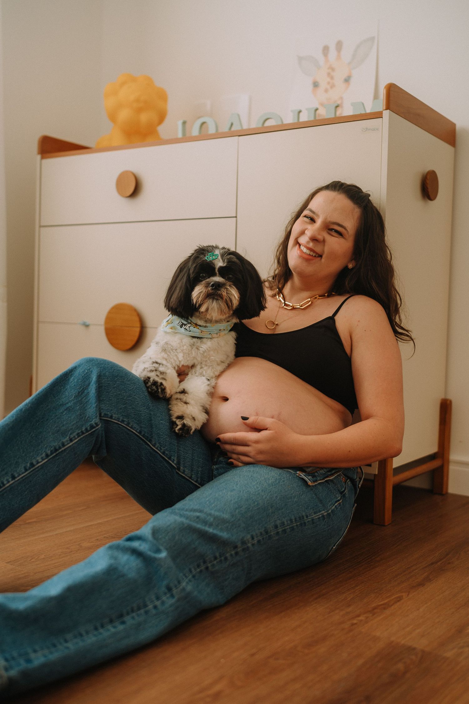 Expecting mother sits on floor with small Shih Tzu dog against white dresser.