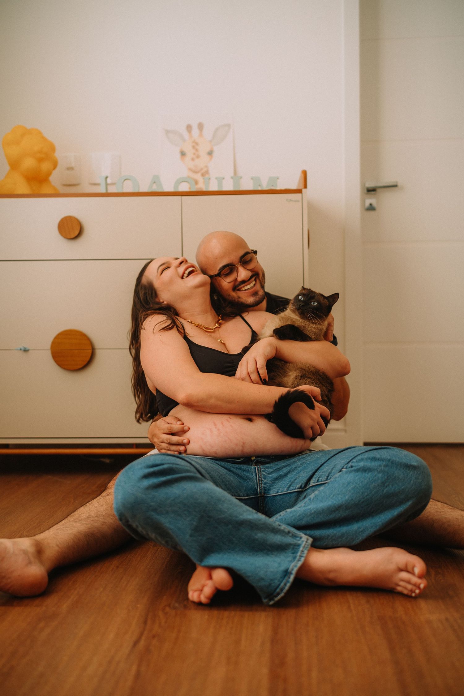 Joyful couple sitting on floor with cat, laughing during maternity photoshoot.