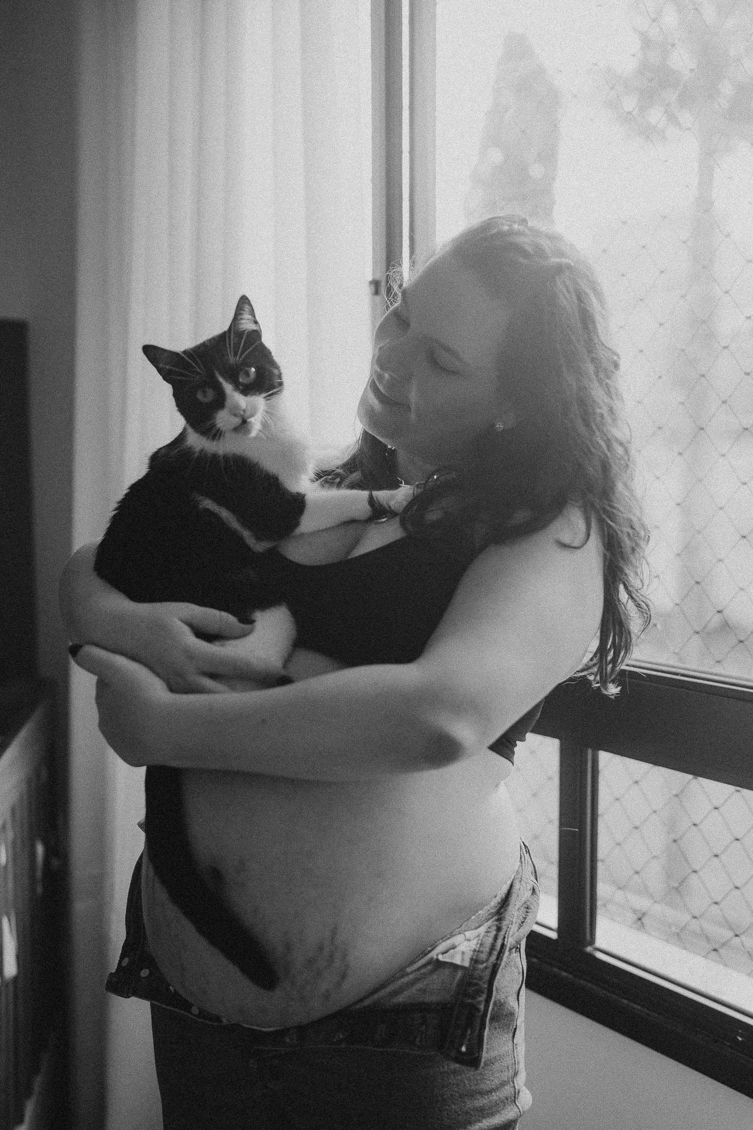 A black and white photo of a pregnant woman holding a black and white cat by a window.