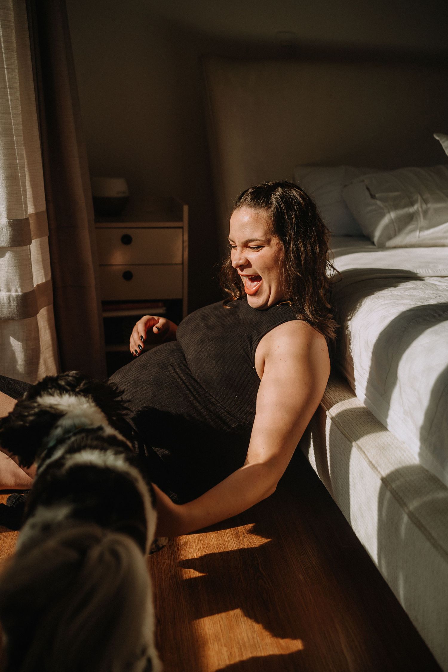 Candid maternity photo showing expectant mother lounging on hardwood floor next to bed in natural lighting.