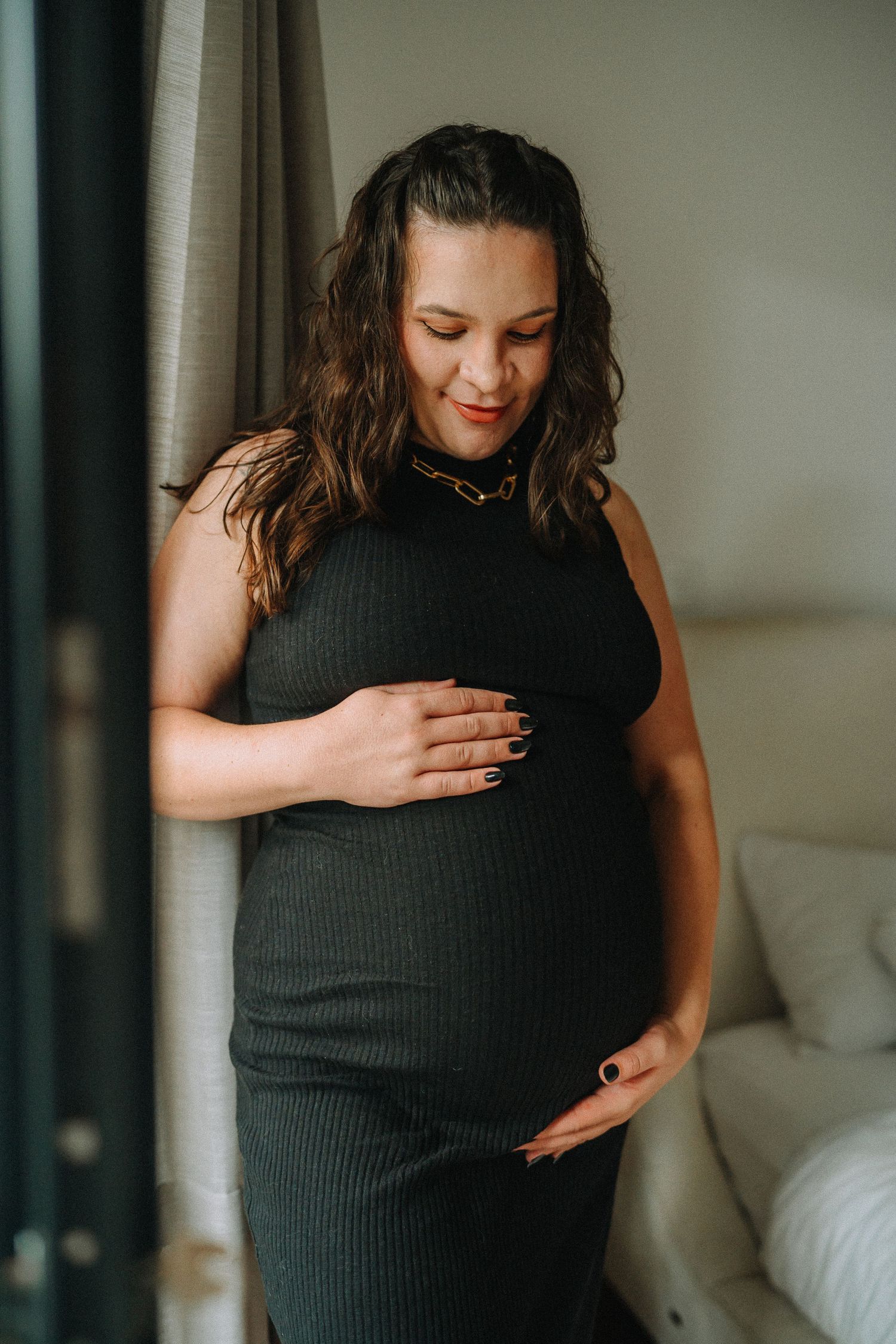Intimate maternity portrait with woman in black ribbed dress by window.
