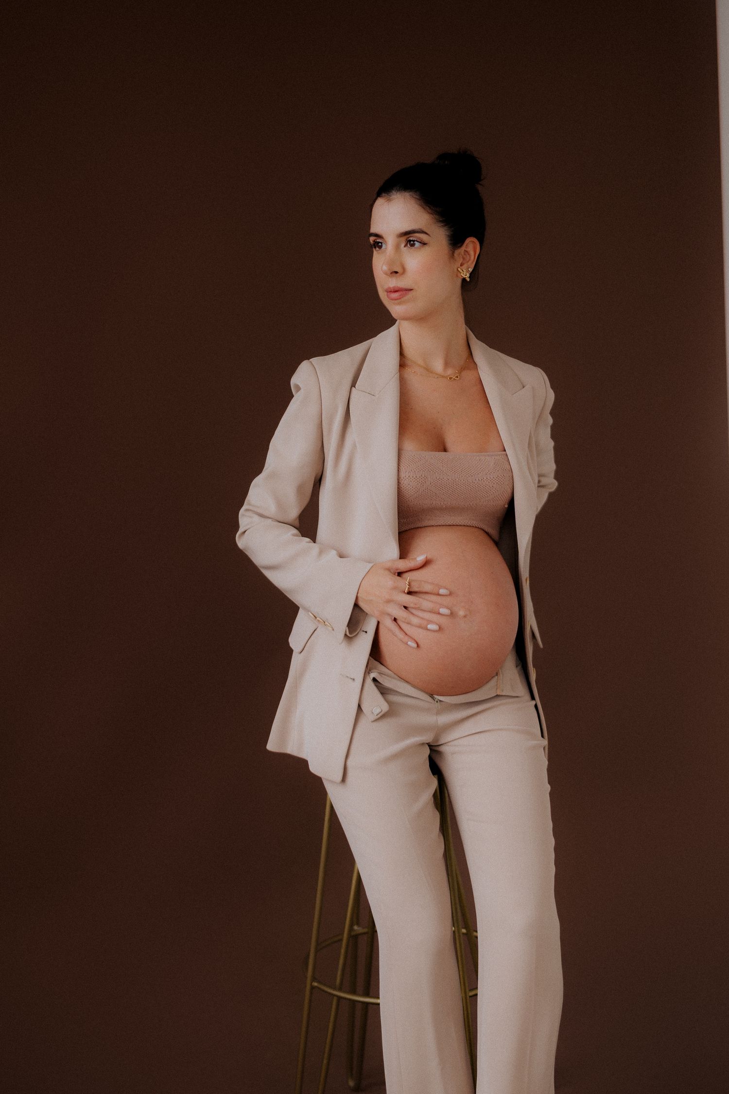 Expectant woman in beige pantsuit poses confidently against dark background.
