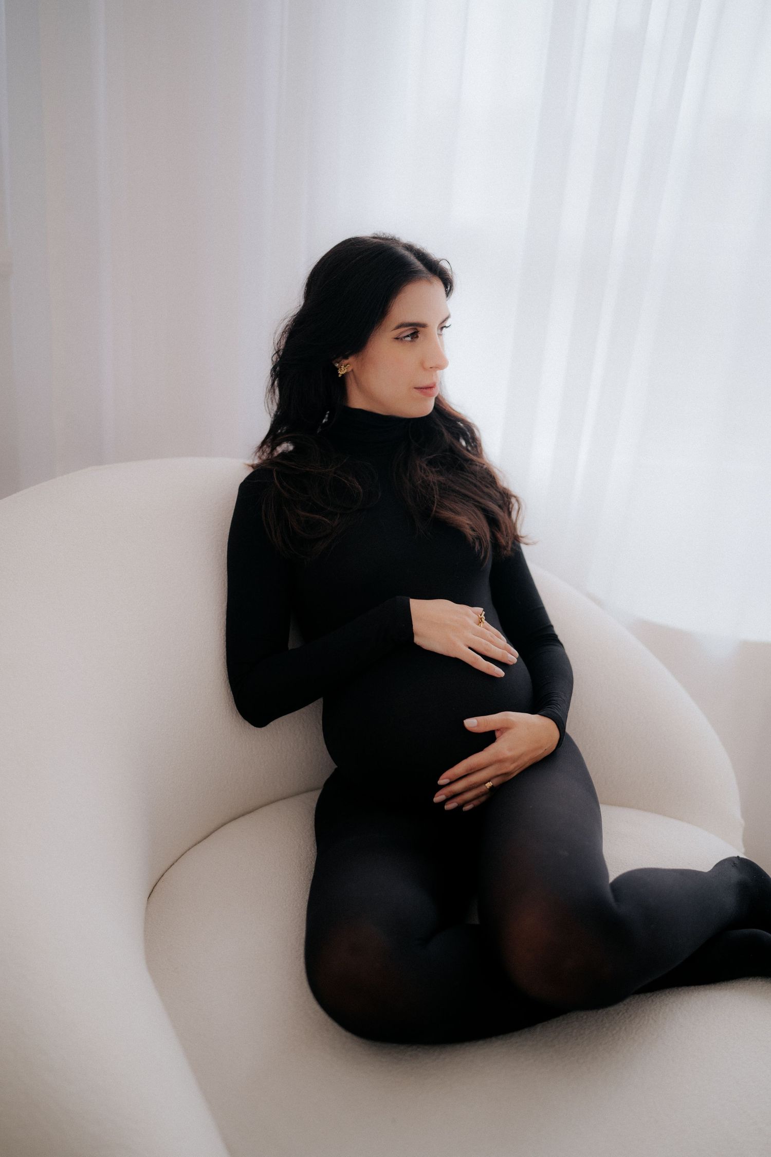 Expectant mother in black dress rests peacefully on white couch during maternity photoshoot.