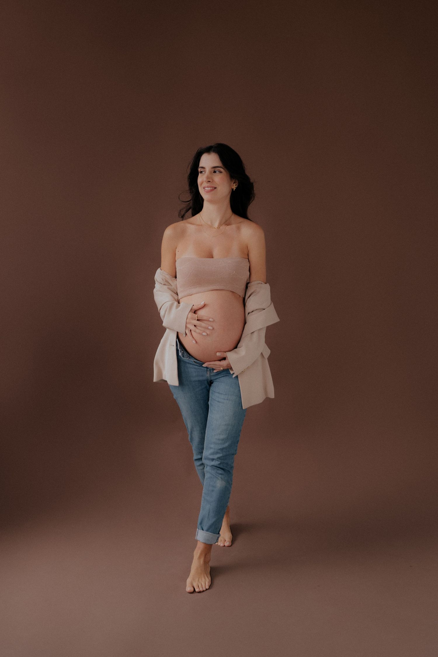 Barefoot pregnant woman in jeans and beige cardigan poses against brown backdrop.