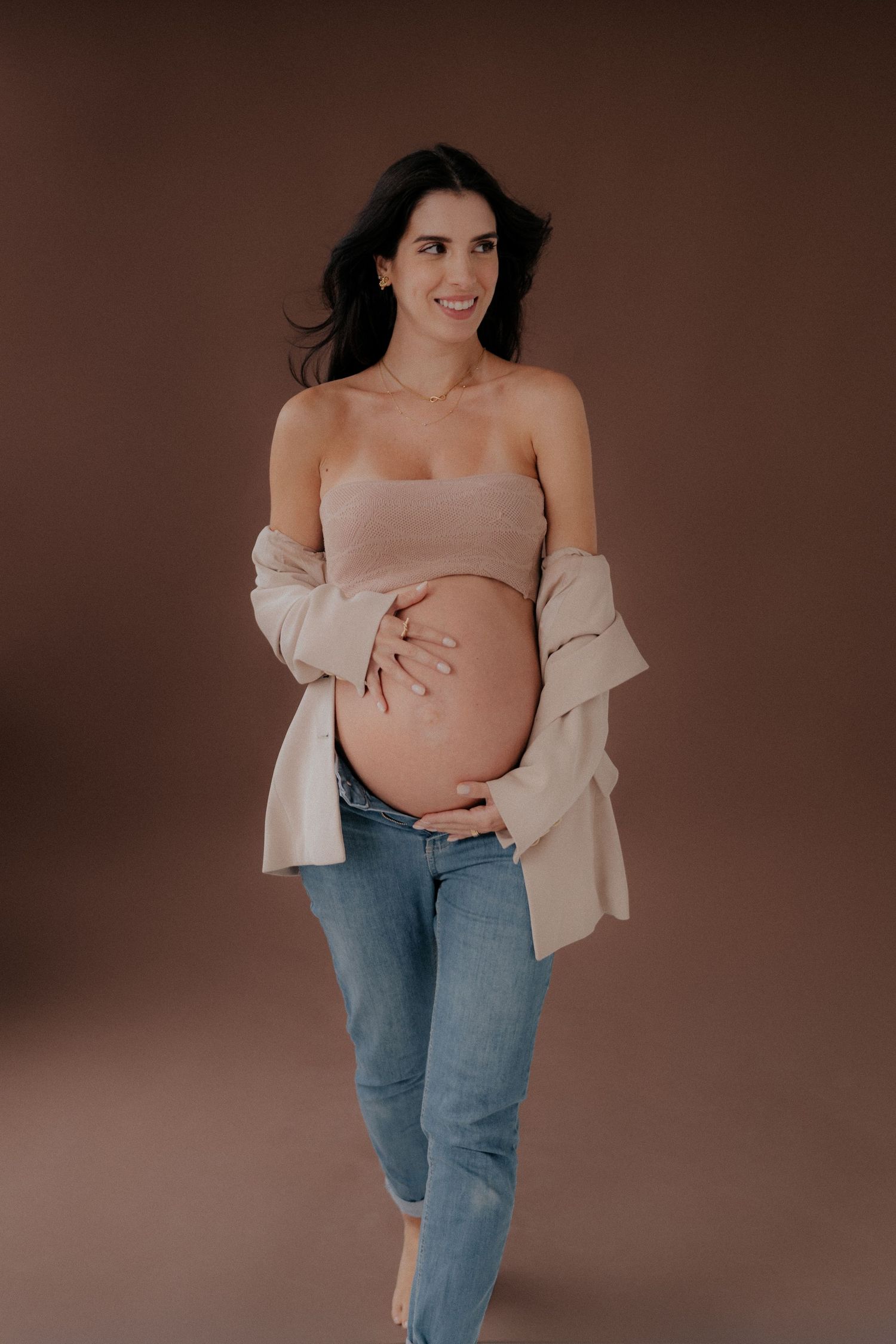 Maternity photo of woman in casual denim and beige cardigan against brown background.
