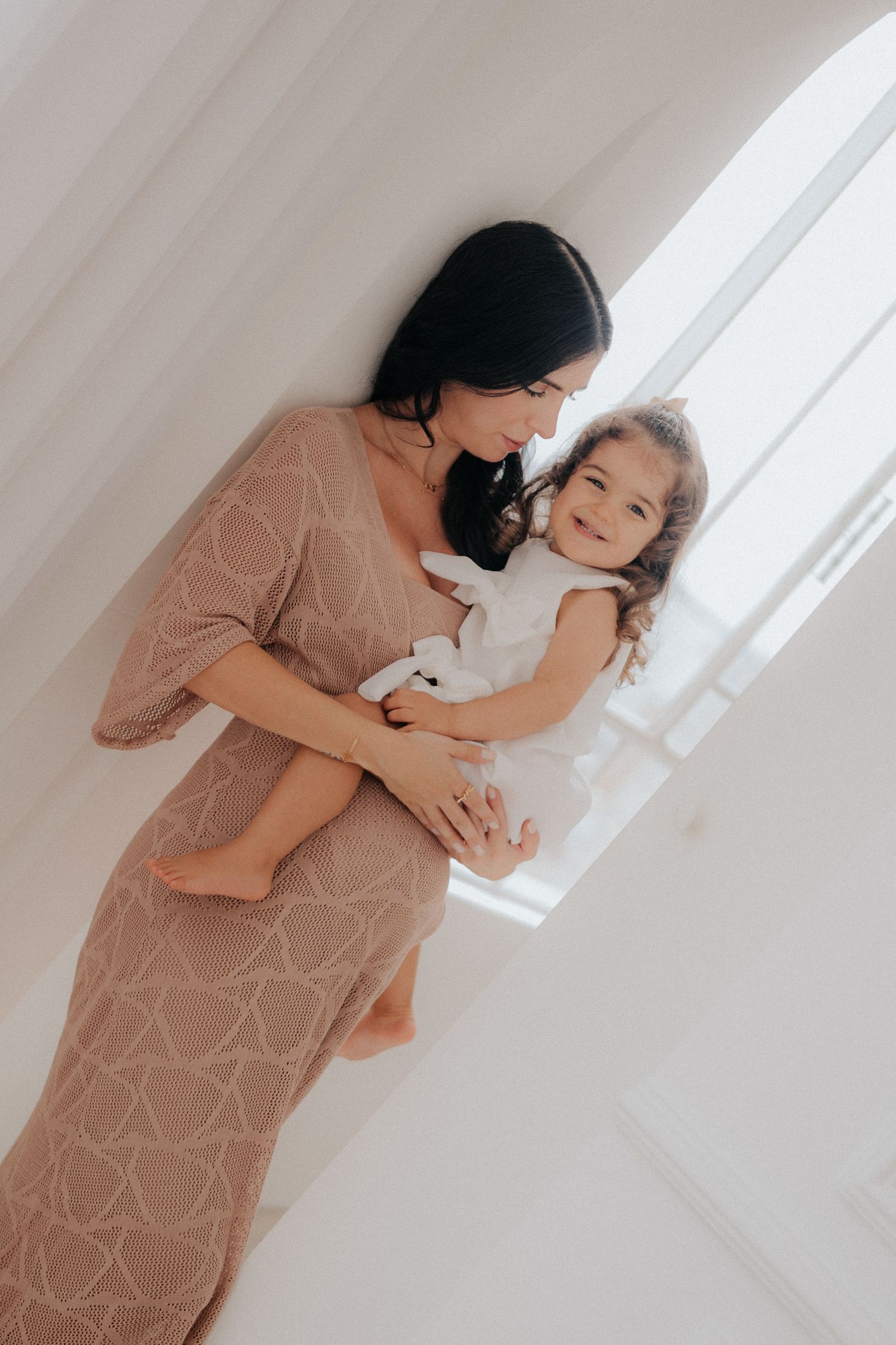 Mother and child share a tender moment near a window wearing matching light-colored outfits.