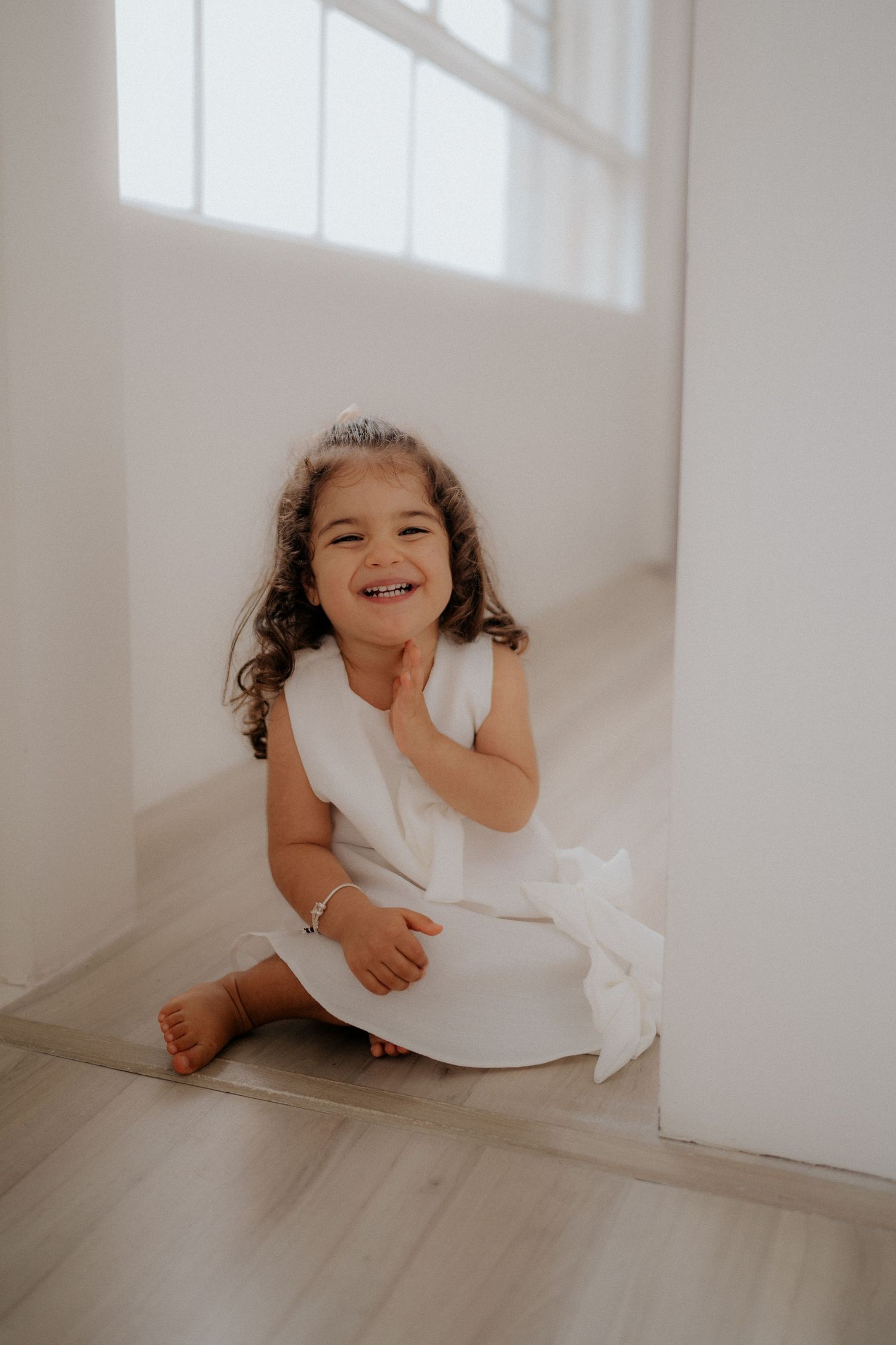 Young child in white dress sits on floor laughing joyfully by window.