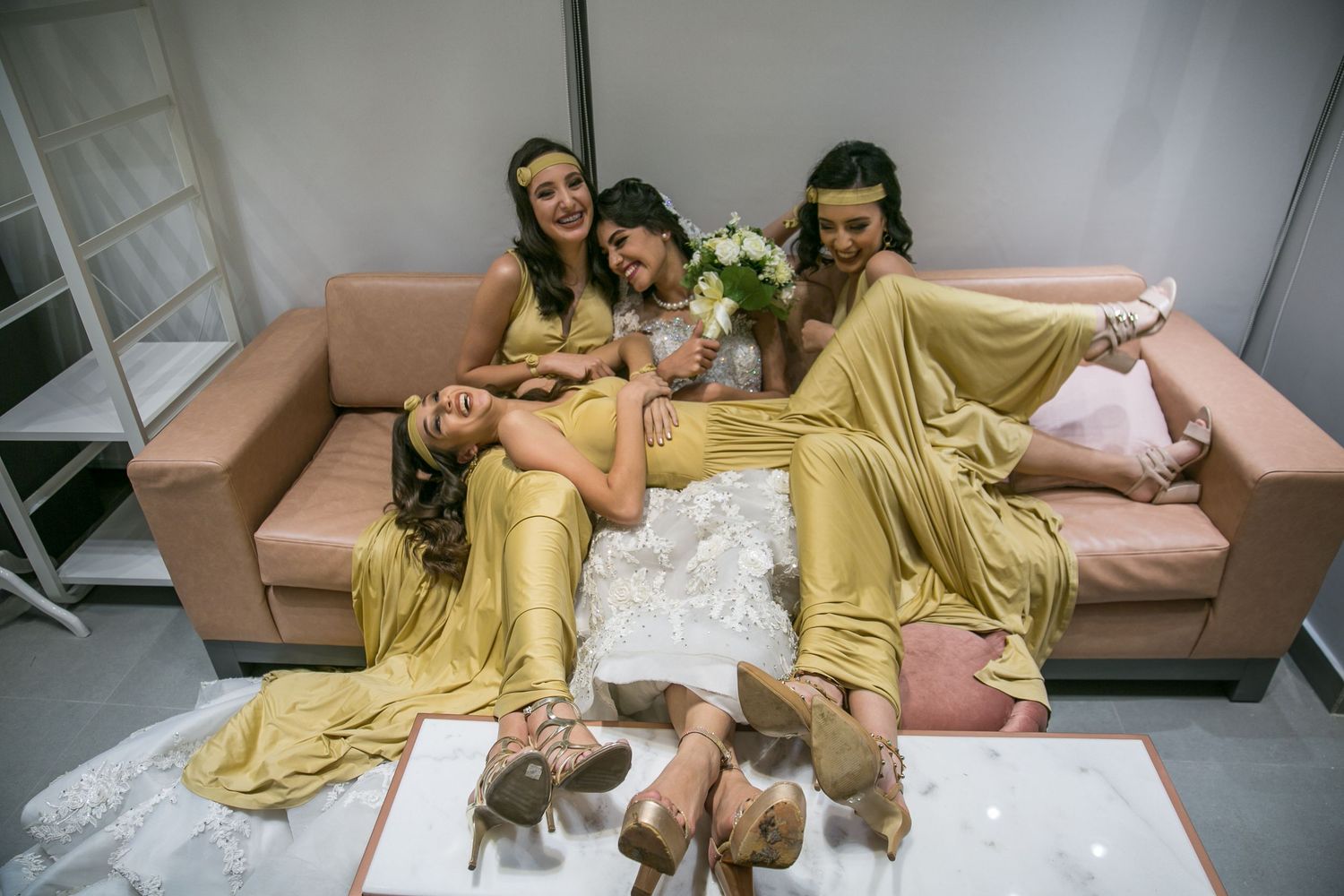 Bride Maids in mustard dresses gathered around the bride fun posing on a light colored couch wearing matching yellow outfits.