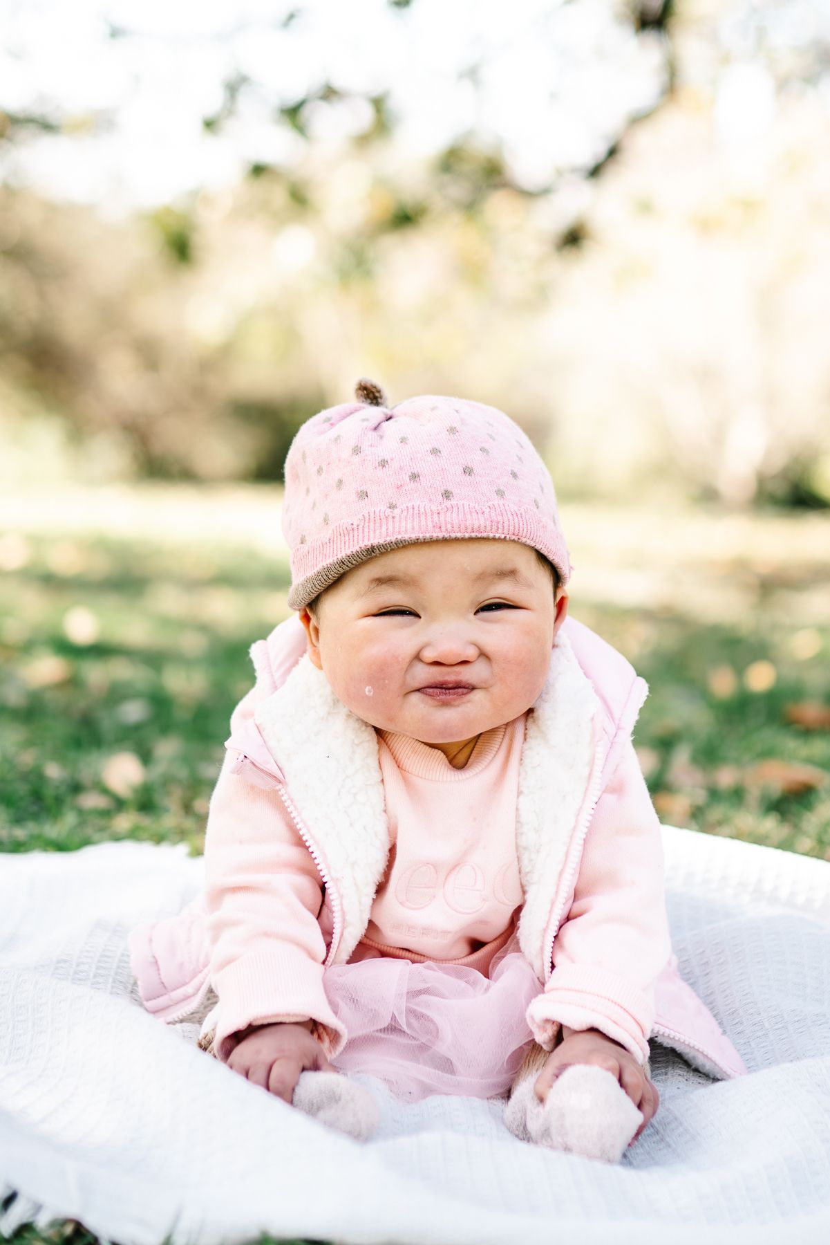 Baby wearing pink outfit and beanie hat smiles while sitting on white blanket outdoors taken during her Melbourne cake smash photo session.