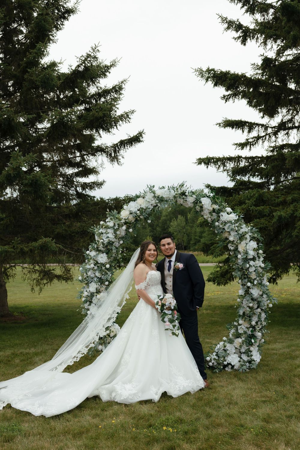 Wedding couple poses beneath a floral arch outdoors with evergreen trees and flowing white dress in the background.