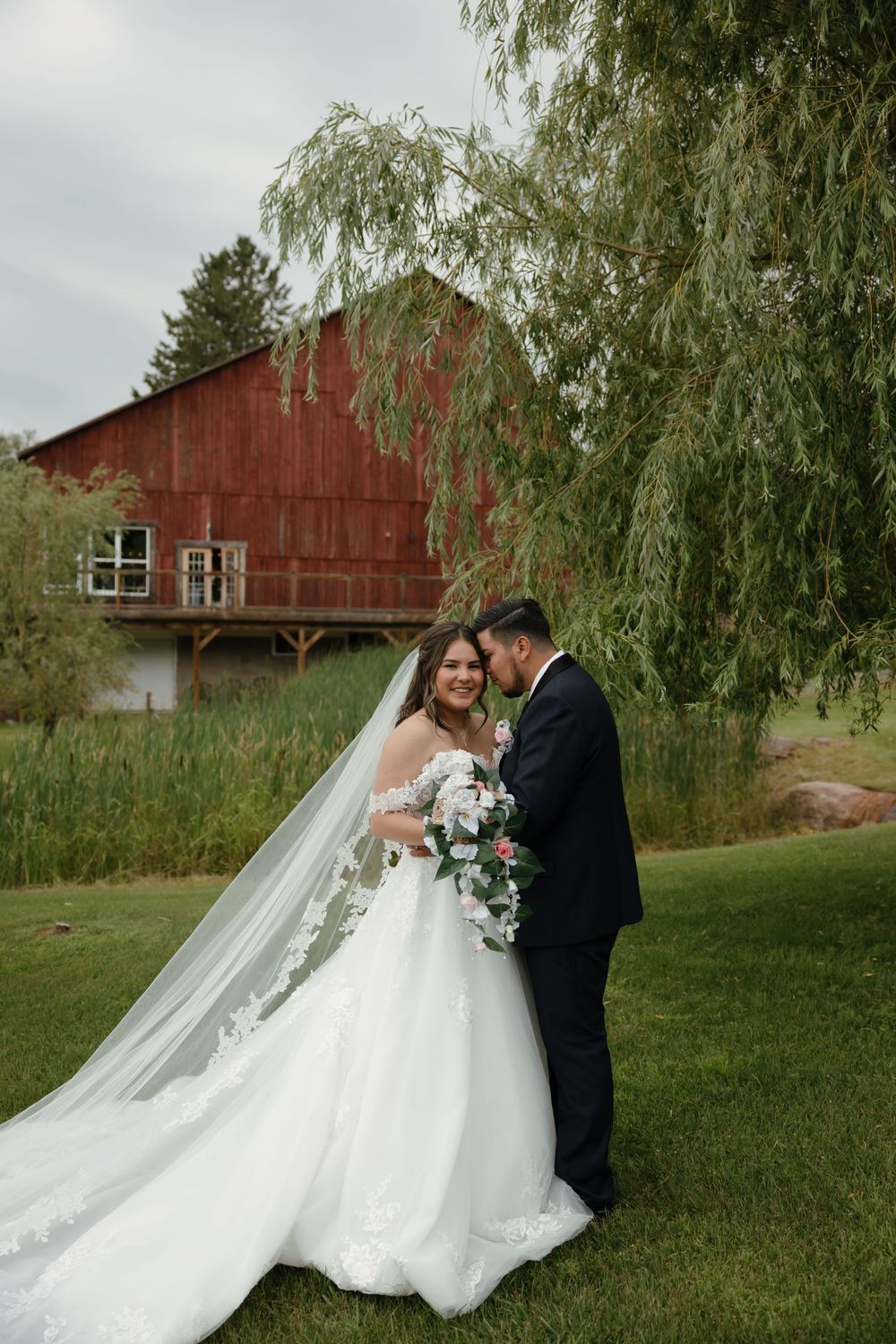 A romantic outdoor wedding photoshoot on a lawn with a rustic red barn in the background.