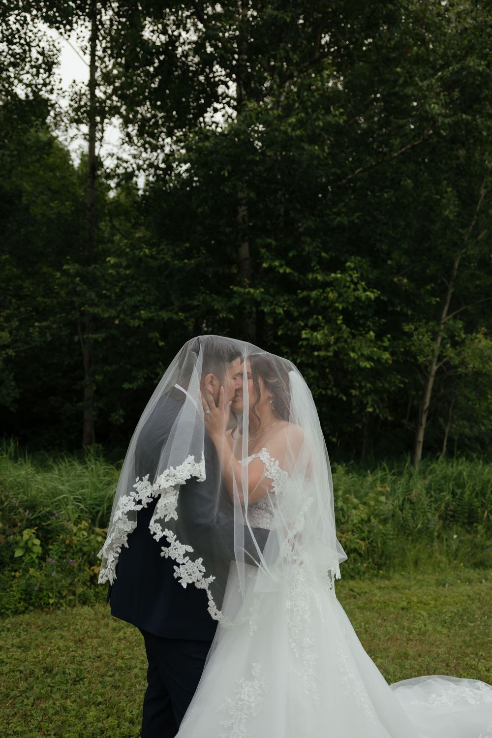 A bride and groom share an intimate moment under a delicate white veil against a backdrop of lush green foliage.