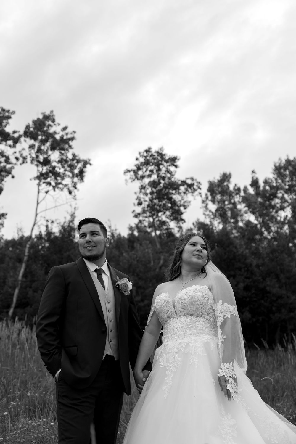 A black and white wedding portrait showing a couple in formal attire standing together in a field with trees in background.