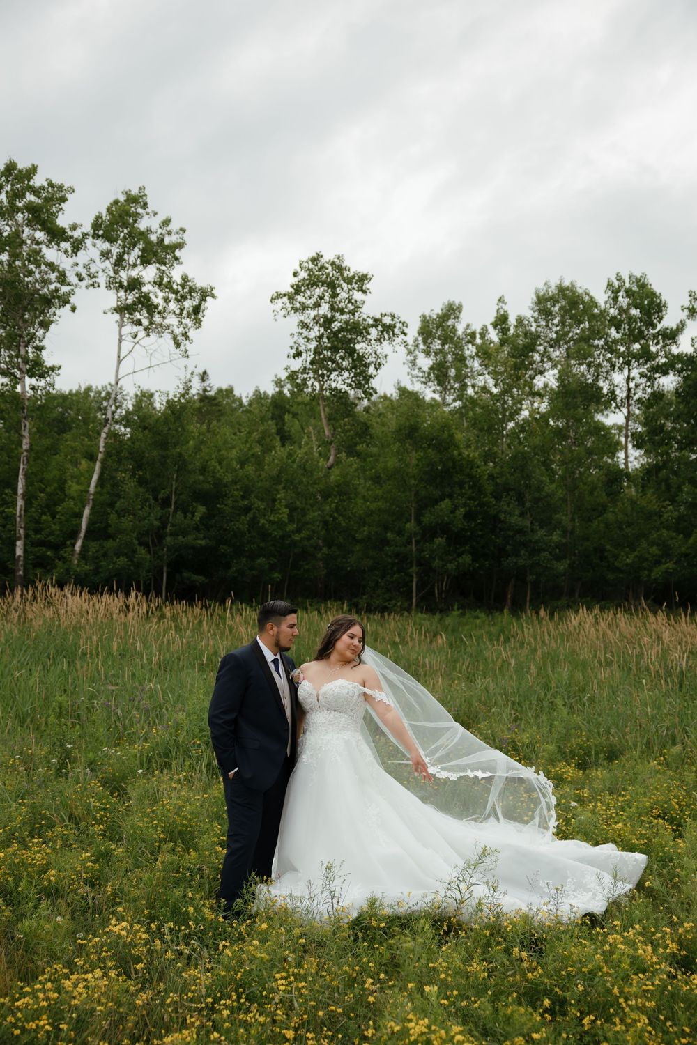 A couple in wedding attire poses gracefully in a field of yellow wildflowers as the bride's veil flows in the breeze.