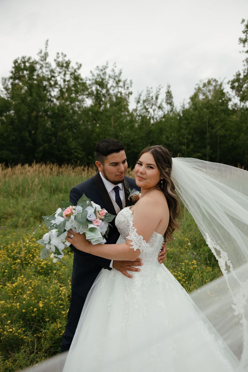 A romantic wedding couple embraces in a field of yellow wildflowers with a flowing white veil trailing behind them.