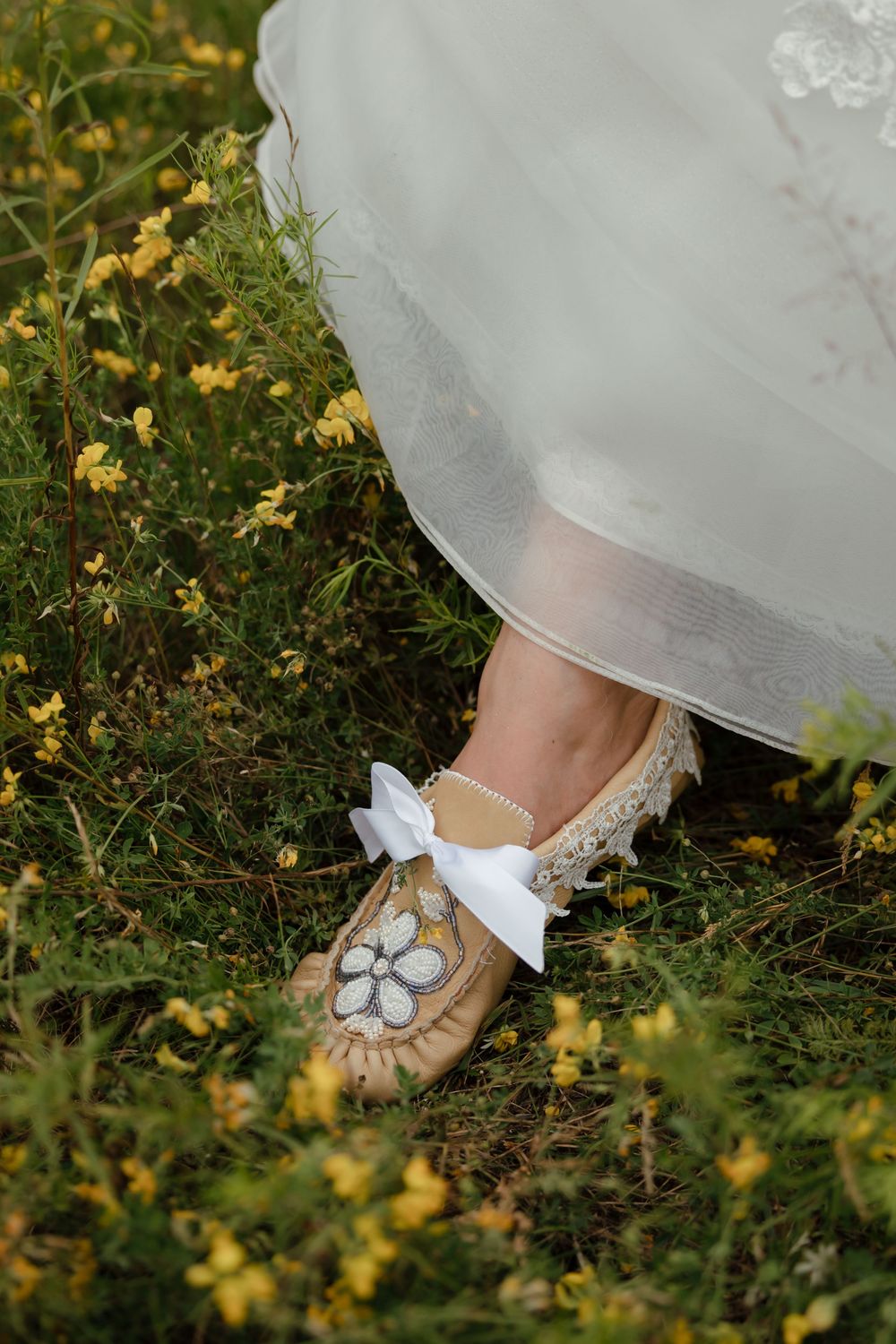 Close-up of a bride's delicate lace shoes peeking out from under her white wedding dress among yellow flowers.
