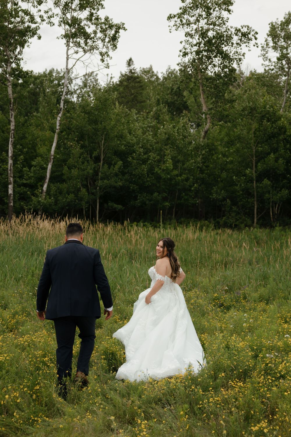 Couple in wedding attire walking through a meadow of wildflowers with birch trees and forest in the background.
