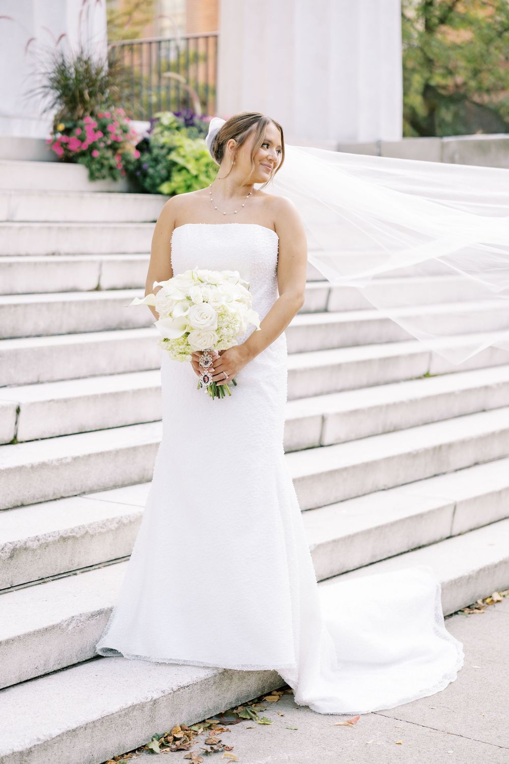 Elegant bride in flowing white gown descending white stone steps in a romantic outdoor setting with greenery.