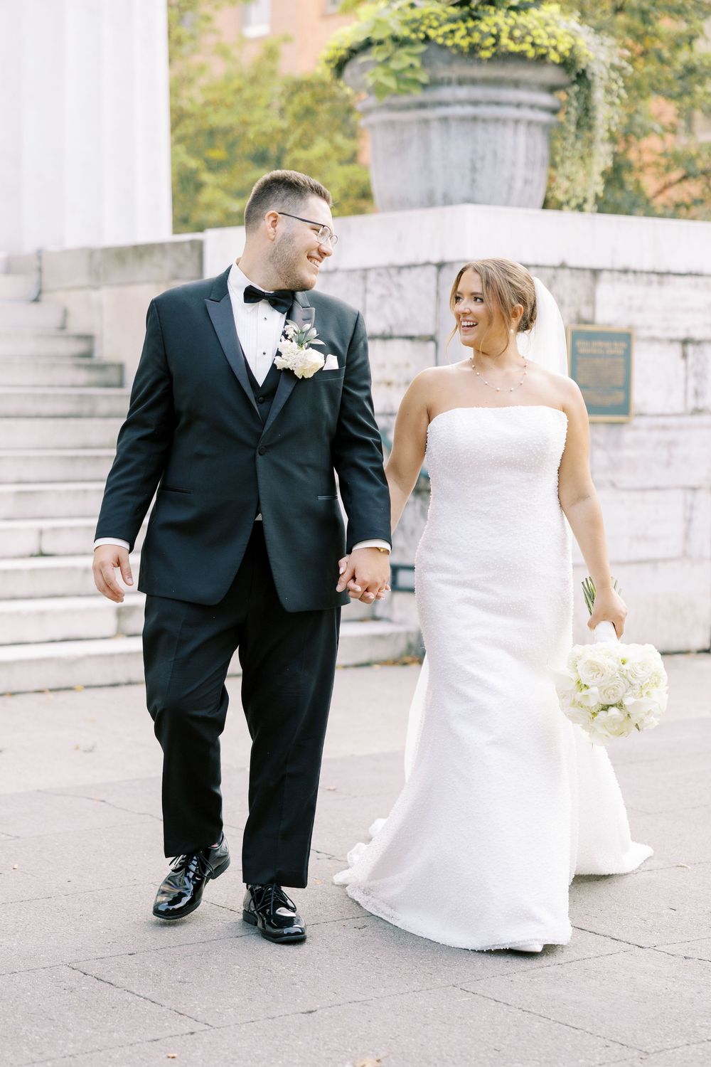 A bride and groom in formal wedding attire stroll hand in hand along a stone pathway.