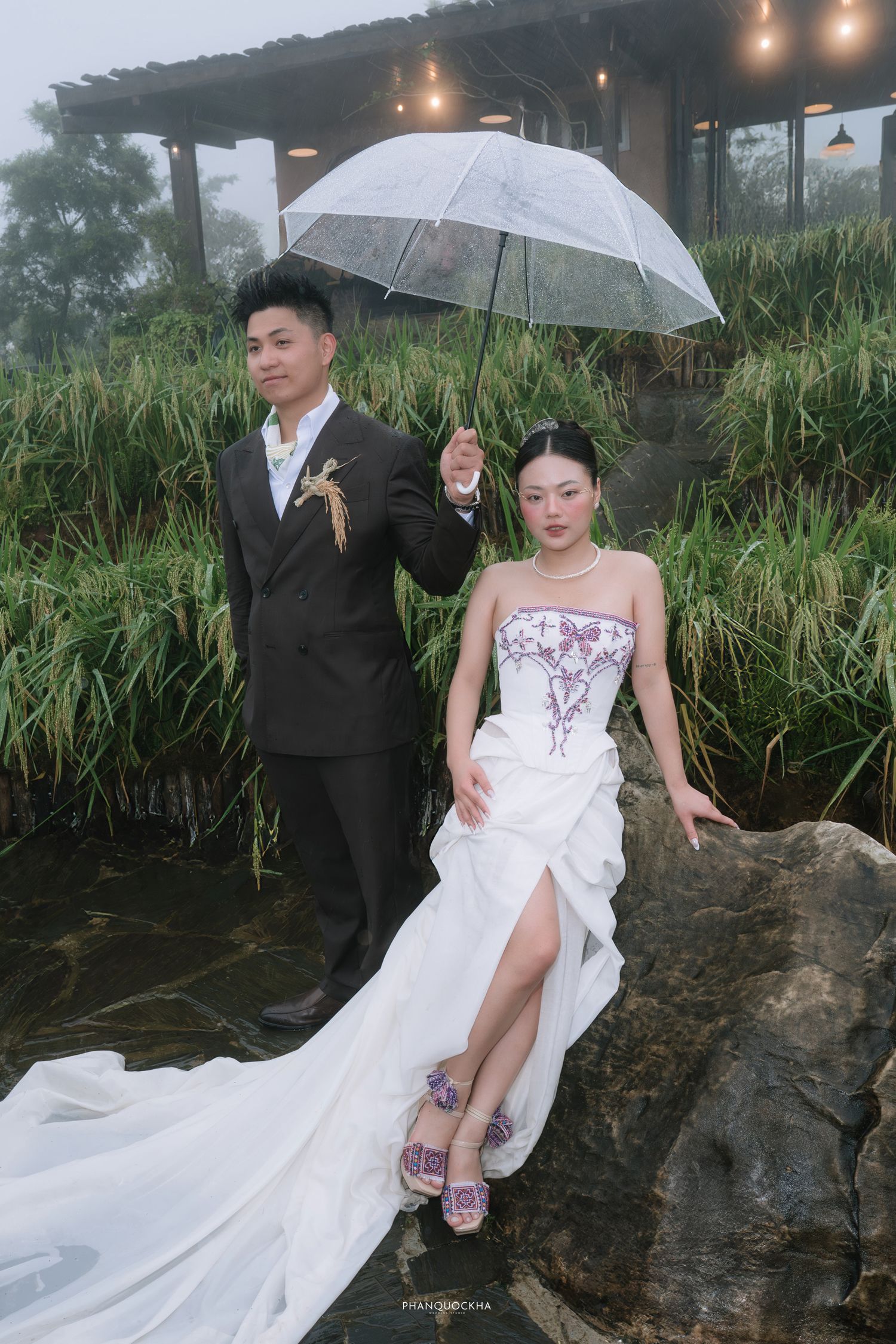 Groom holds umbrella over bride in embroidered white gown seated on rocks by misty garden pond.