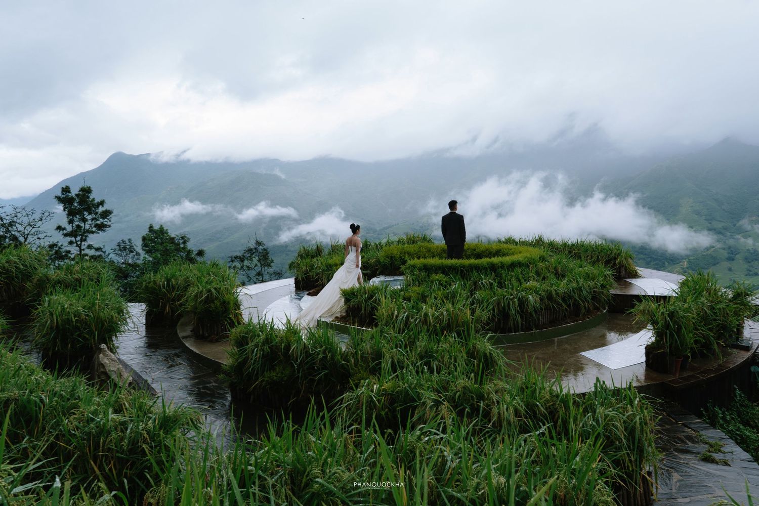 Bride and groom stand on curved rice terrace path surrounded by green fields and misty mountains.