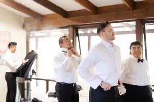 Groomsmen in formal black and white attire chat and interact while getting ready in a rustic indoor venue.