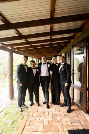 Groomsmen in black tuxedos pose playfully by leaning against a covered walkway with brick flooring.