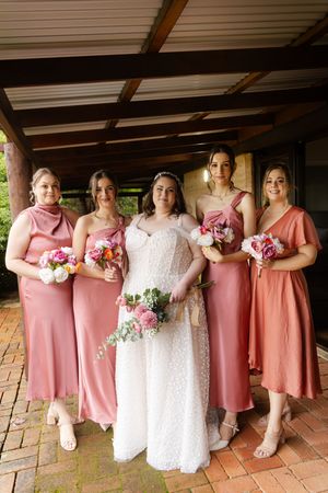 Bridesmaids in coral pink dresses pose with the bride in her white gown under a covered outdoor patio.