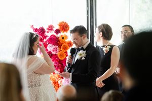 A couple exchanges vows during a wedding ceremony in front of a vibrant orange and pink floral backdrop.