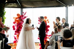 Modern indoor wedding vows exchange with bright red flower decorations framing glass windows.