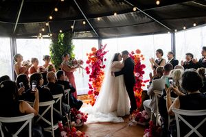 Wedding guests in a decorative reception venue with white chairs and red floral arrangements along the wall.