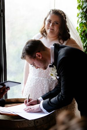 Wedding ceremony paperwork being signed at a wooden table near bright windows.