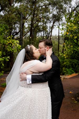 wedding couple share a kiss on a tree-lined path outdoors while wearing formal attire.