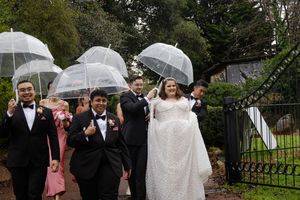Wedding party members walk through rain holding clear umbrellas for outdoor photos at a garden venue.